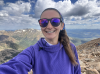 A selfie of Taylor Young. She is at the summit of Mount Elbert in a purple sweatshirt, sunglasses, and a purple-blue headband. There are some fluffy clouds in the background that contrast the cool blues, greens, and browns of the sky and mountains. 