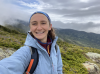 A selfie of Taylor Young. She is somewhere along the Presidential Range of the White Mountains. She's in a blue rain jacket with green, mountains, and clouds in the background.